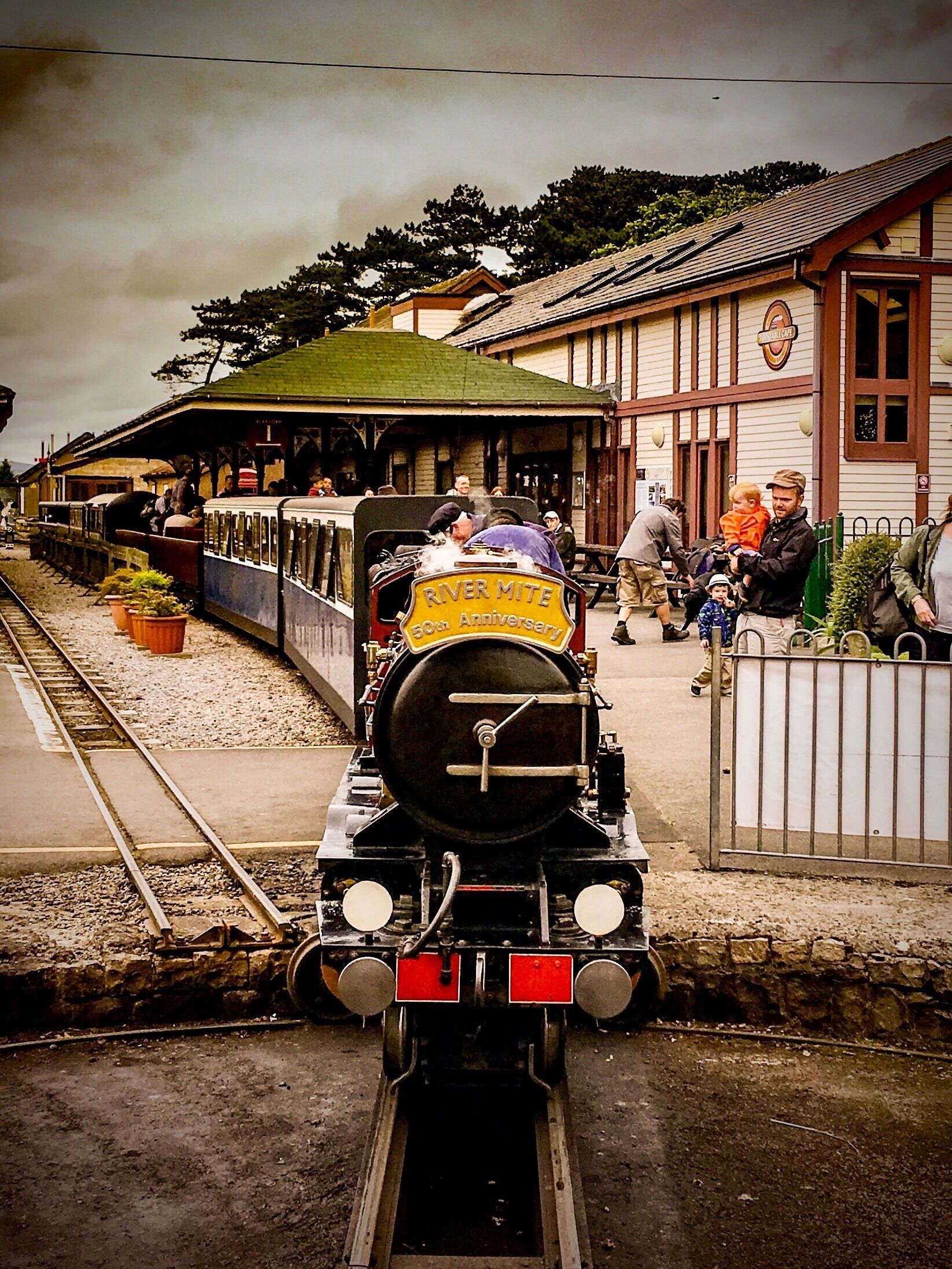 A steam train with a 'River Mite 50th Anniversary' sign, passengers, and a station with a green roof and wooden buildings.