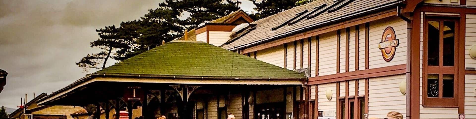 A steam train with a 'River Mite 50th Anniversary' sign, passengers, and a station with a green roof and wooden buildings.