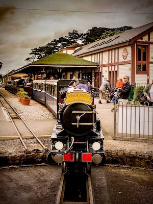 Un tren de vapor con un letrero que dice 'River Mite 50th Anniversary', pasajeros y una estación con techo verde y edificios de madera.