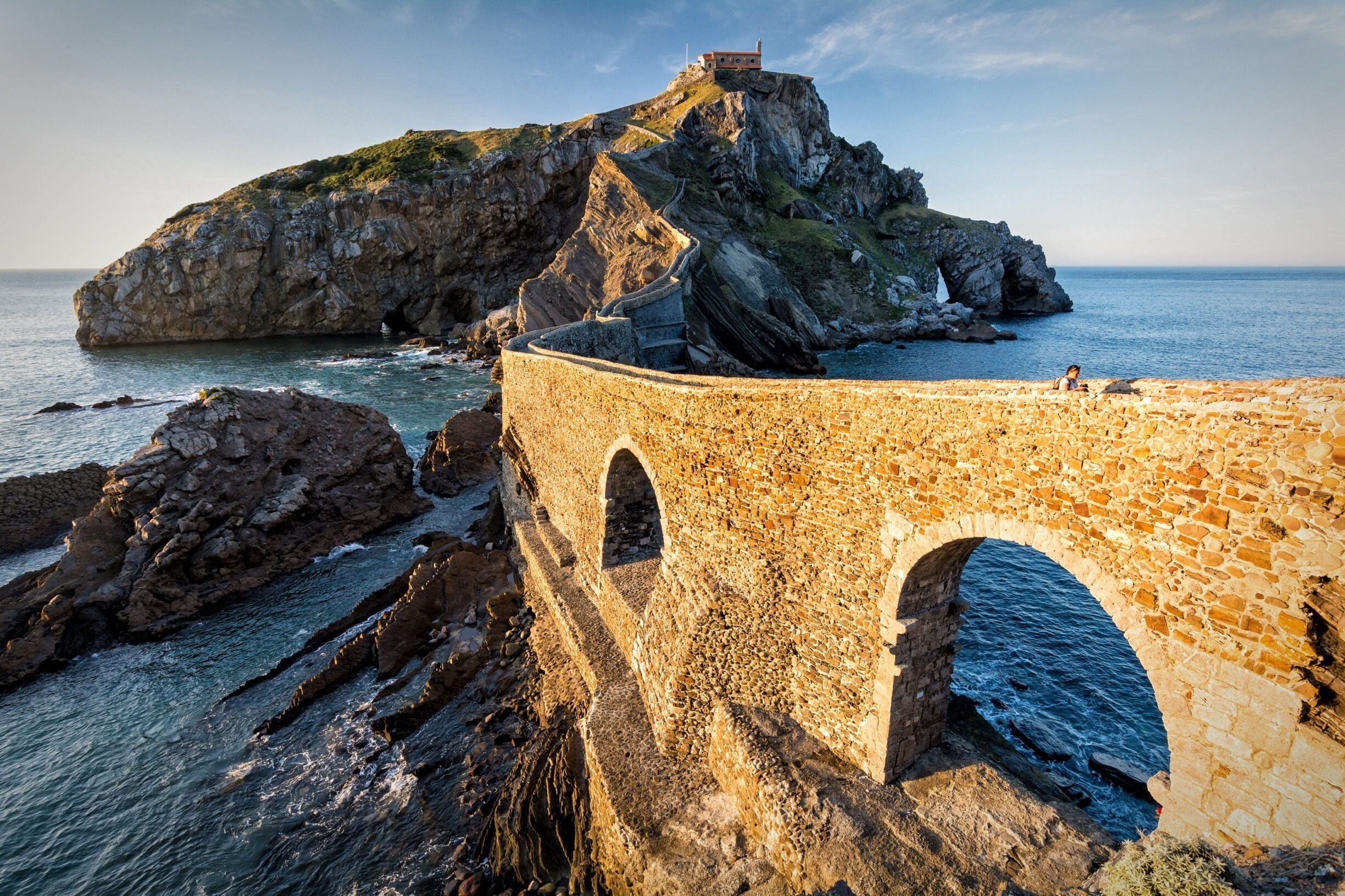 Un puente de piedra con arcos, costa rocosa y un faro en una colina.