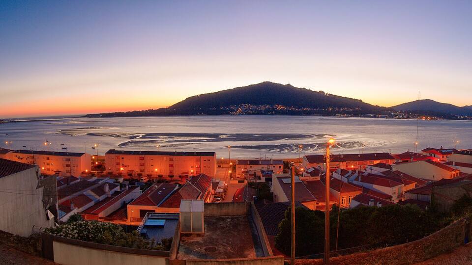 A coastal town at dusk with a large body of water, a hill in the background, and buildings with red roofs.