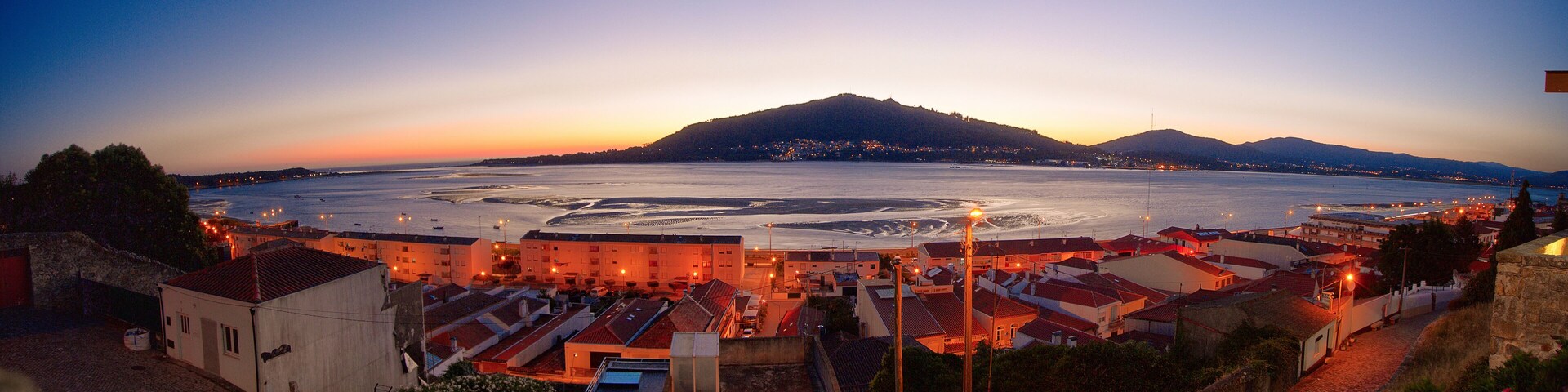 A coastal town at dusk with a large body of water, a hill in the background, and buildings with red roofs.
