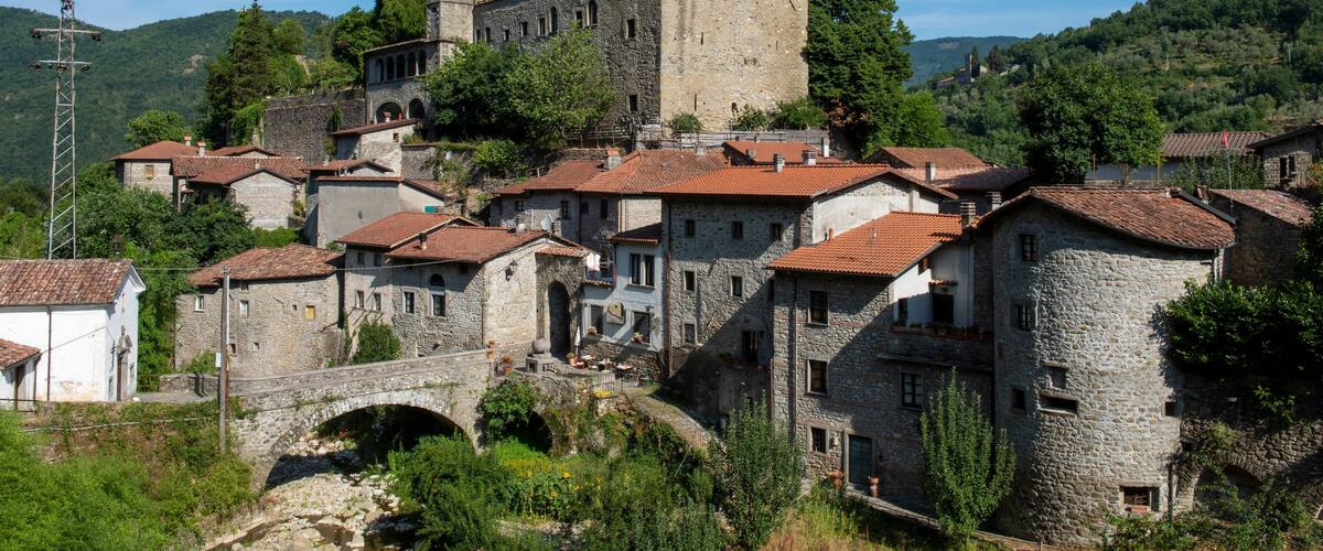 A medieval castle atop a hill, surrounded by stone buildings with red-tiled roofs, a stone bridge, and lush greenery.
