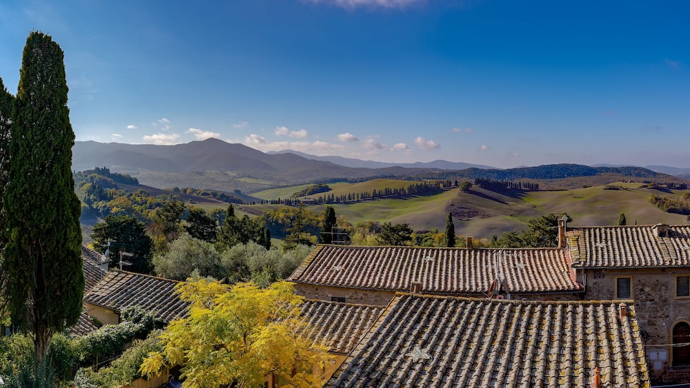 Un paysage rural caractérisé par des collines douces, un grand cyprès et des bâtiments traditionnels aux toits de tuiles.