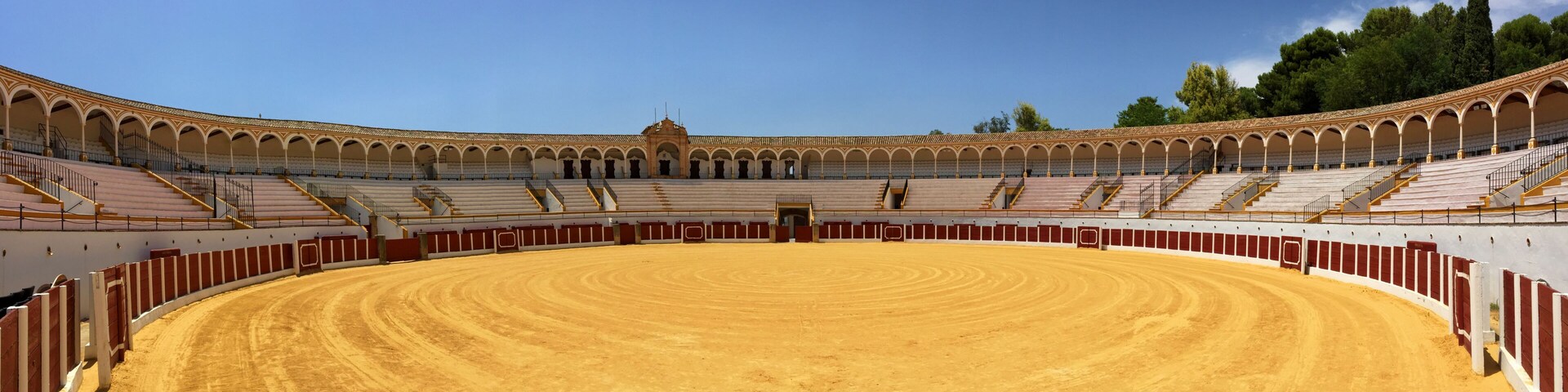 A large, empty bullring with a sandy floor and red and white walls.