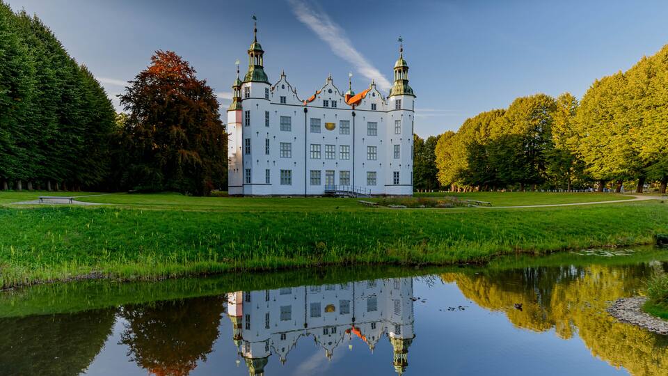 A white castle with green roofs and multiple towers, surrounded by a pond reflecting the structure, and a park with trees and grass.