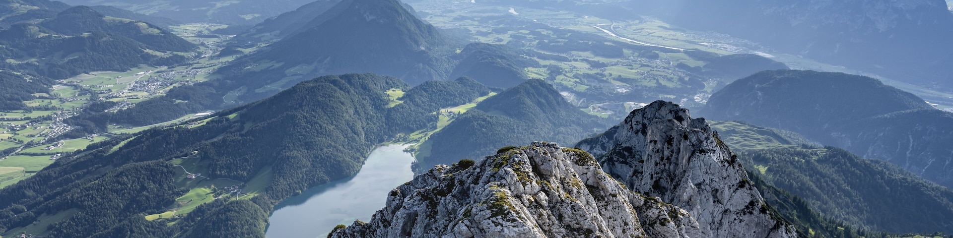 A mountainous landscape with a prominent rocky peak in the foreground and a valley with a river in the background.