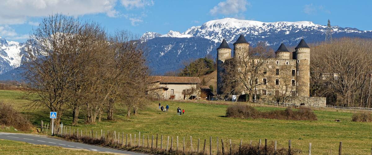 A castle with three towers, a mountain range with snow, and a clear blue sky.