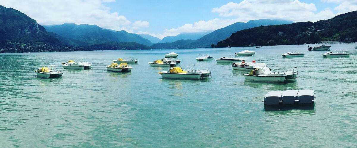 Un lac paisible où flottent plusieurs bateaux, entouré de montagnes et surplombé d’un ciel bleu clair.