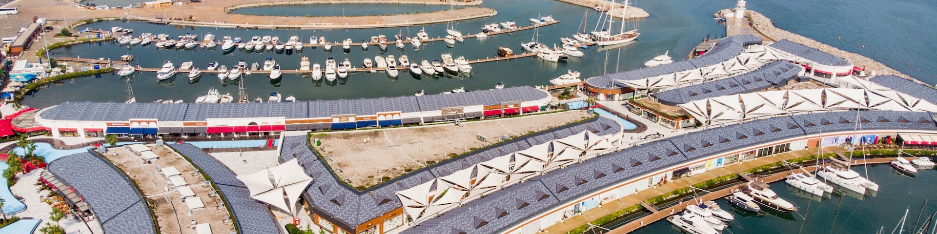 Vista aérea de un puerto deportivo con numerosas embarcaciones amarradas, un tren pasando y un paisaje urbano costero al fondo.