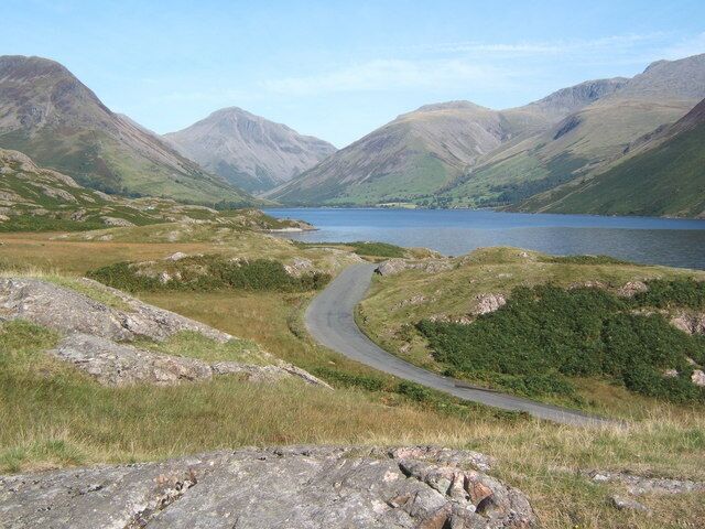 Eine kurvenreiche Straße durch eine gebirgige Landschaft, im Hintergrund ein See.