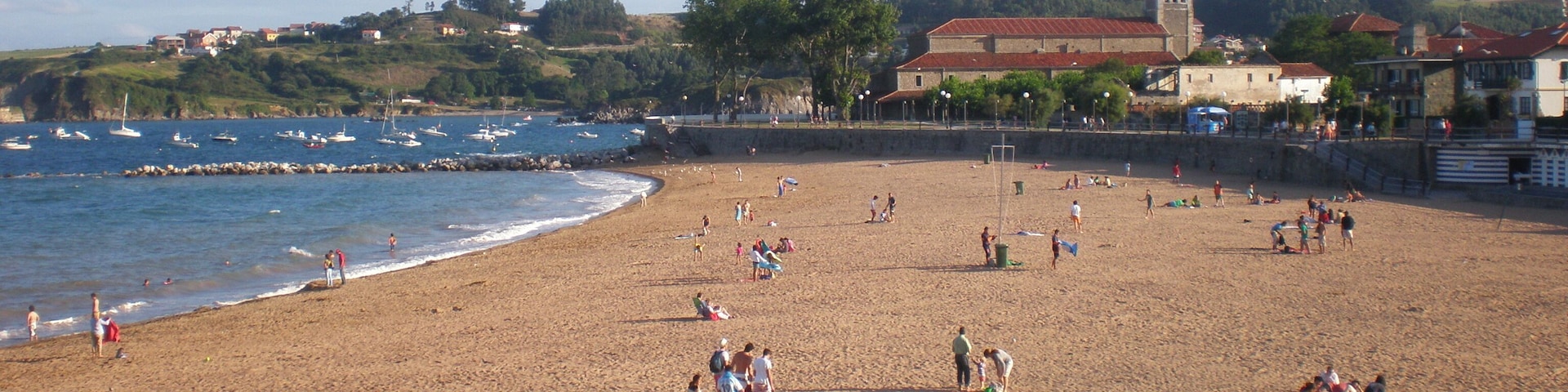 Uma praia arenosa com pessoas, barcos e uma igreja ao fundo.