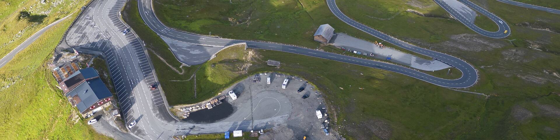 Aerial view of winding roads cutting through verdant hills near a cluster of buildings, a testament to human ingenuity amidst nature's grandeur, Bruck an der Großglocknerstraße, Salzburg, Austria.