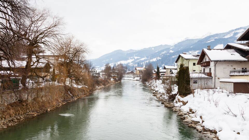 A river flowing through a snow-covered village with mountains in the background.