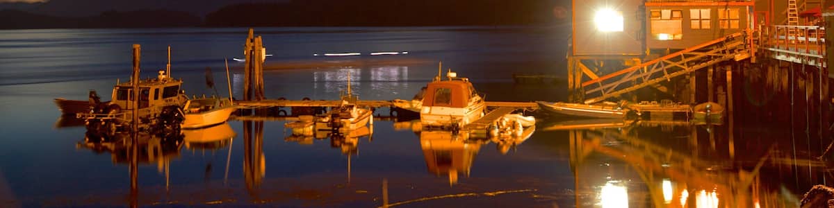 Un lac paisible au crépuscule, avec des bateaux amarrés, une jetée en bois et un bâtiment illuminé, le tout sur fond de montagnes.