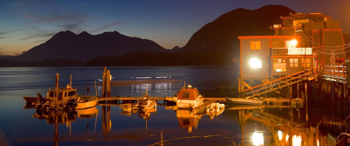 A calm lake at dusk with boats docked, a wooden pier, and a brightly lit building with mountains in the background.