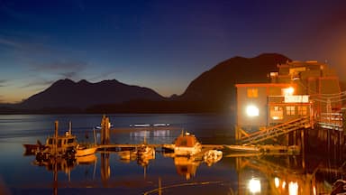 A calm lake at dusk with boats docked, a wooden pier, and a brightly lit building with mountains in the background.