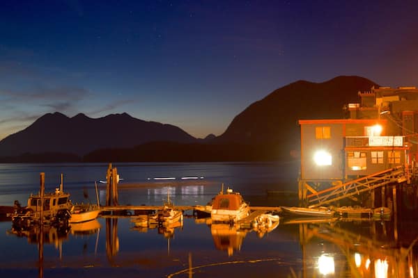 Un lac paisible au crépuscule, avec des bateaux amarrés, une jetée en bois et un bâtiment illuminé, le tout sur fond de montagnes.