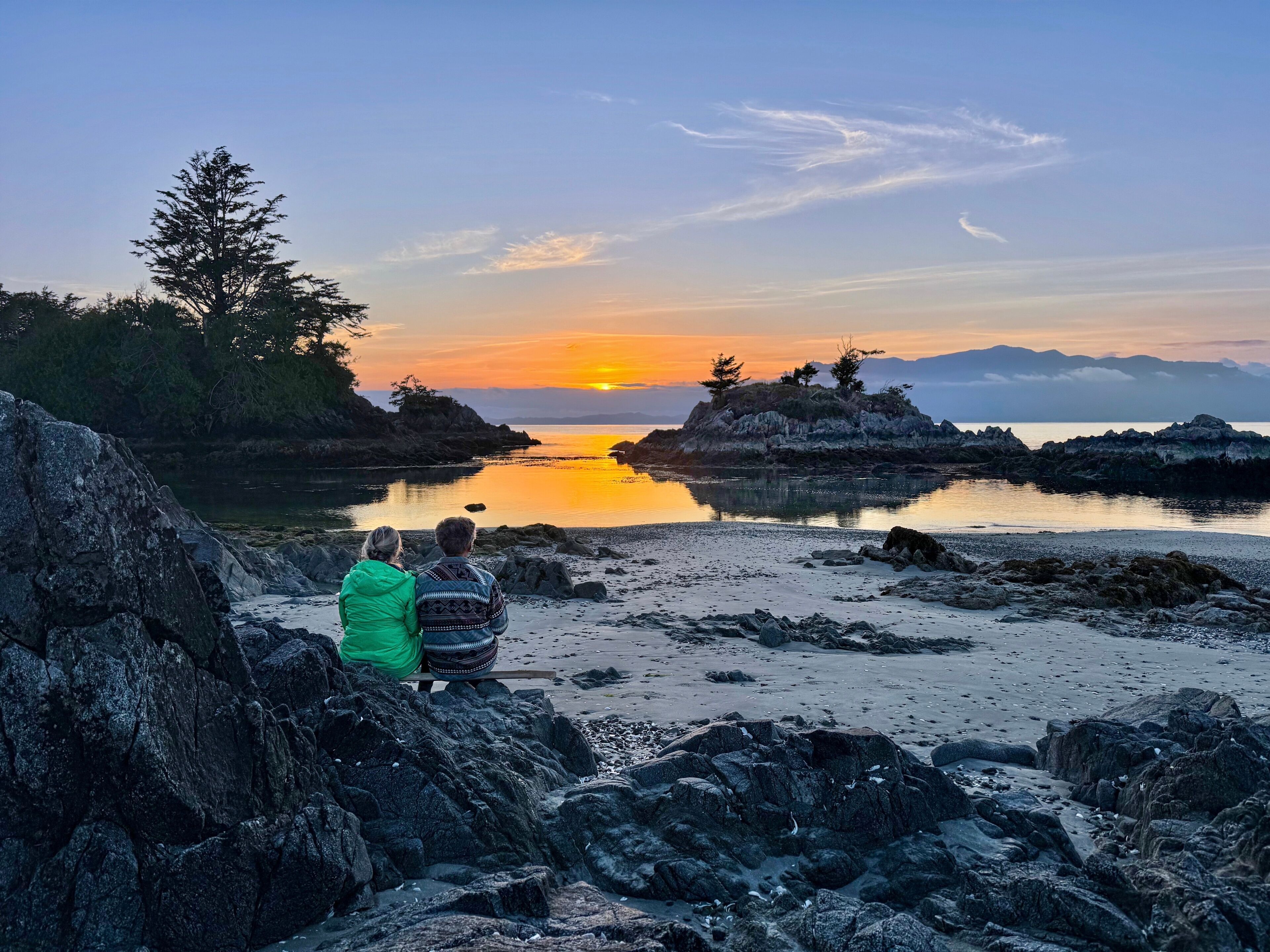 A rocky shoreline with a sunset over water and distant mountains.