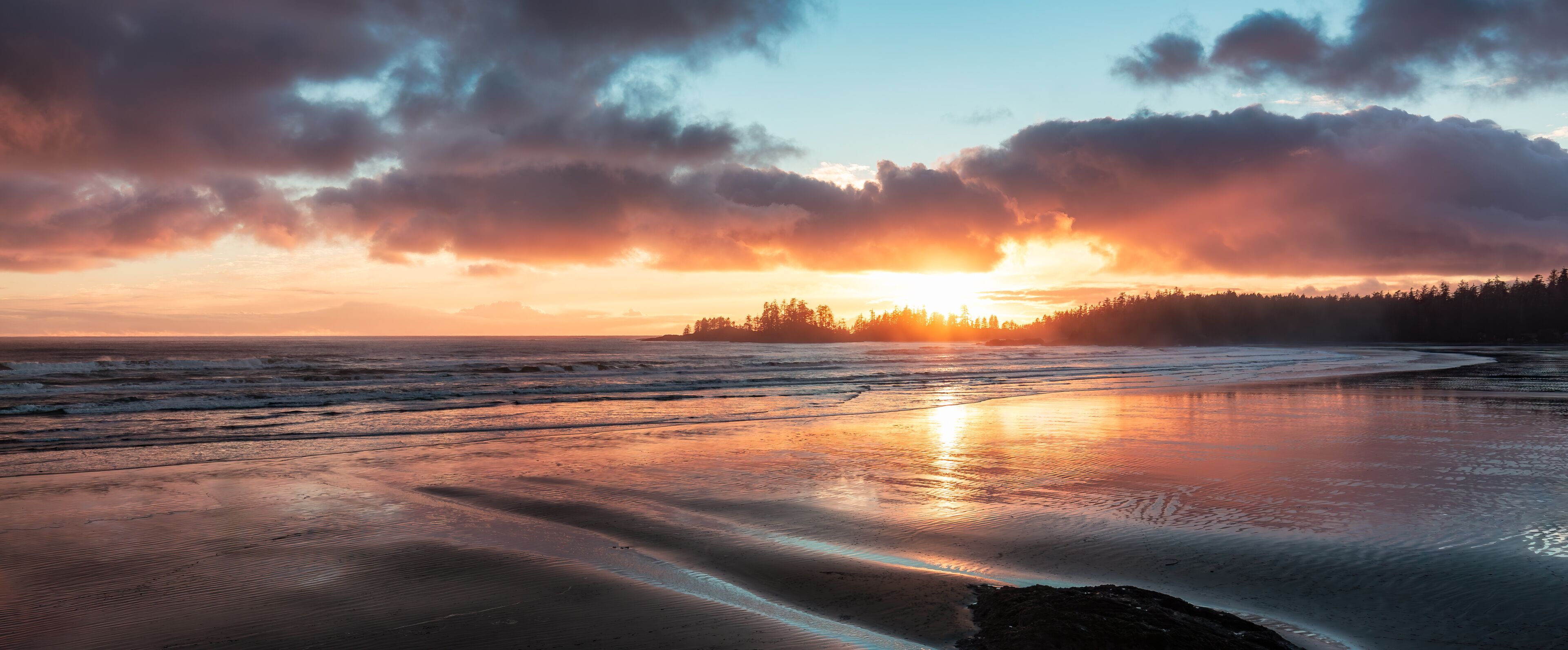 Una playa al atardecer con olas, un bosque en el fondo y un cielo nublado.