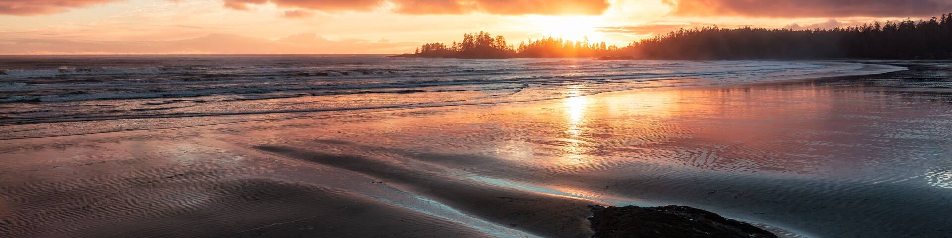 Una playa al atardecer con olas, un bosque en el fondo y un cielo nublado.