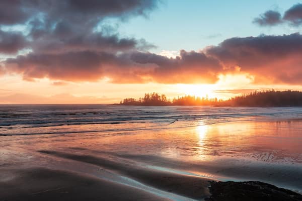 A beach at sunset with waves, a forest in the background, and a cloudy sky.