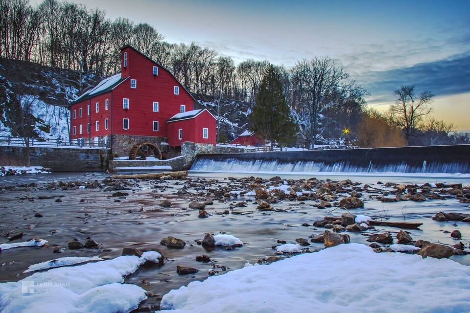 A red watermill by a river with snow-covered ground and trees in the background.