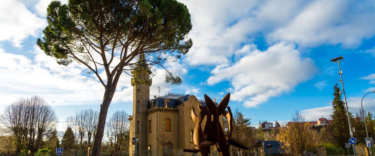 A roundabout with a large tree, a historic building, and a modern sculpture.