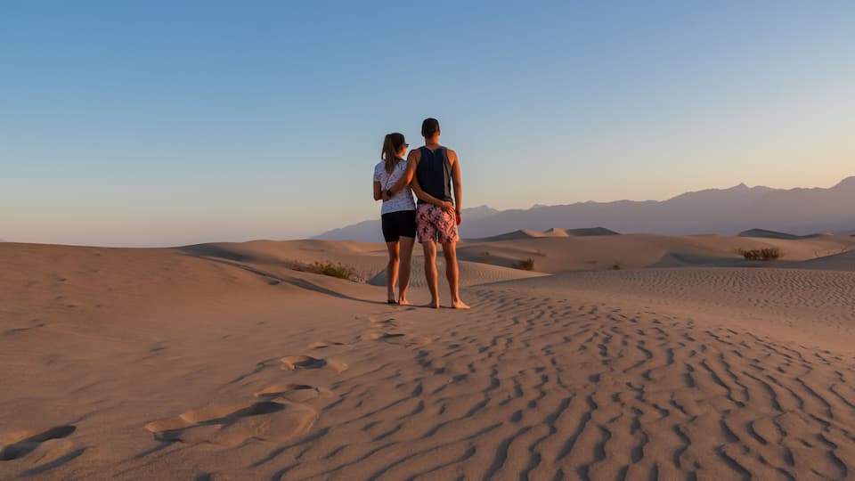 Two people standing on a desert sand dune with mountains in the background.