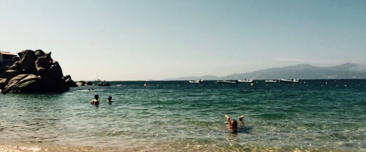 A beach with clear water, people swimming, and boats in the distance.