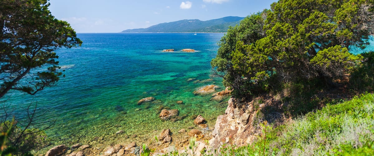 A coastal landscape with clear turquoise waters, rocky shores, and dense green trees.