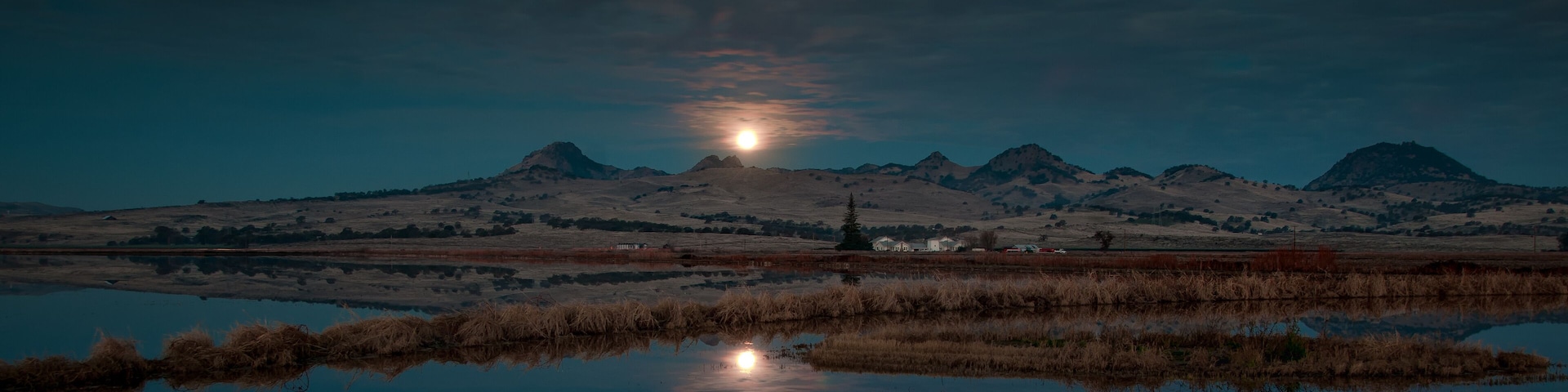 A calm lake reflecting the moon and mountains in the distance.