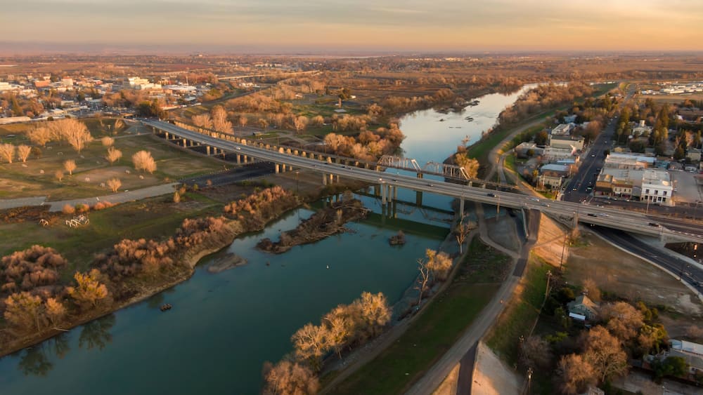 Aerial view of Yuba City, California, USA, at sunset. The Feather River flows under bridges carrying cars and pedestrians. The image shows the city's layout and transportation.