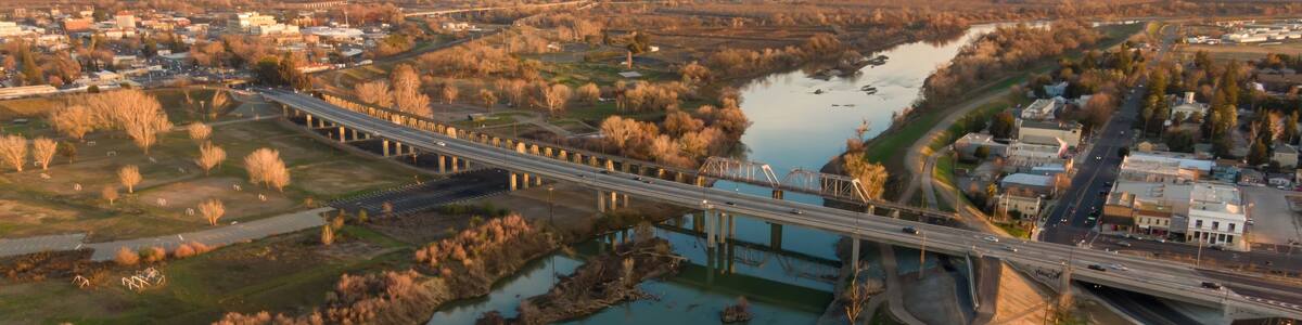 Aerial view of Yuba City, California, USA, at sunset. The Feather River flows under bridges carrying cars and pedestrians. The image shows the city's layout and transportation.