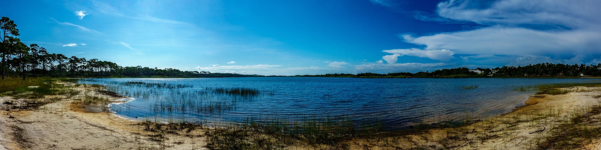 Um lago tranquilo com águas azuis cristalinas, rodeado por margens de areia e árvores verdes densas sob um céu azul vibrante.