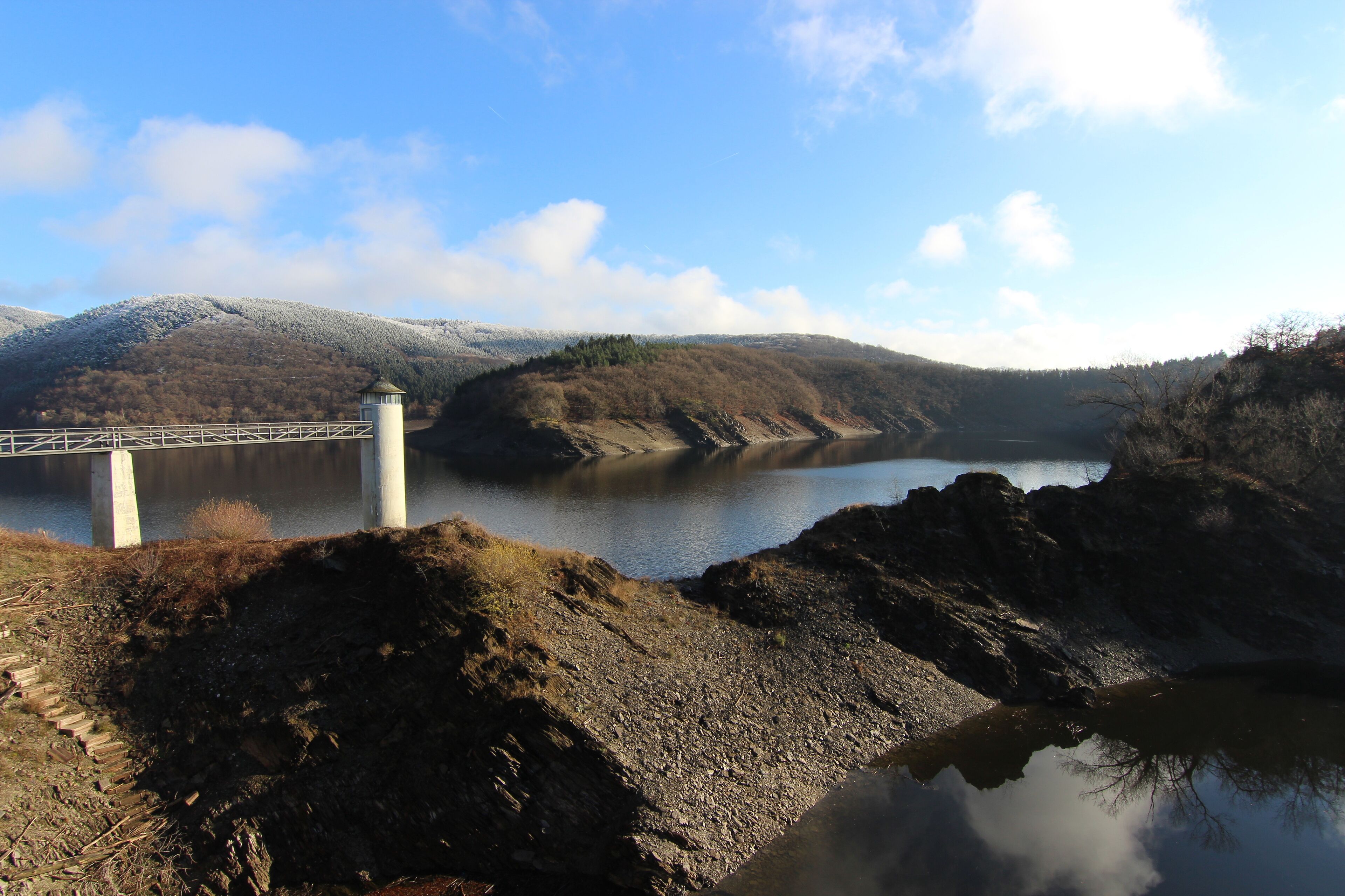 Das Bild zeigt einen Blick von der Urftstaumauer auf den Urftsee im Nationalpark Eifel. – Der Nationalpark repräsentiert die für die nördliche Eifel typischen natürlichen und naturnahen Lebensräume und Lebensgemeinschaften auf bodensauren Standorten von der kollinen bis zur montanen Höhenstufe zwischen 200 m und 600 m. Dies sind insbesondere unterschiedliche Laubwälder, Quellgebiete, Fließgewässer, Offenlandbiotope und Felsbildungen. Die größten Flächenanteile wurden zuvor militärisch verwaltet durch die belgischen Streitkräfte genutzt.