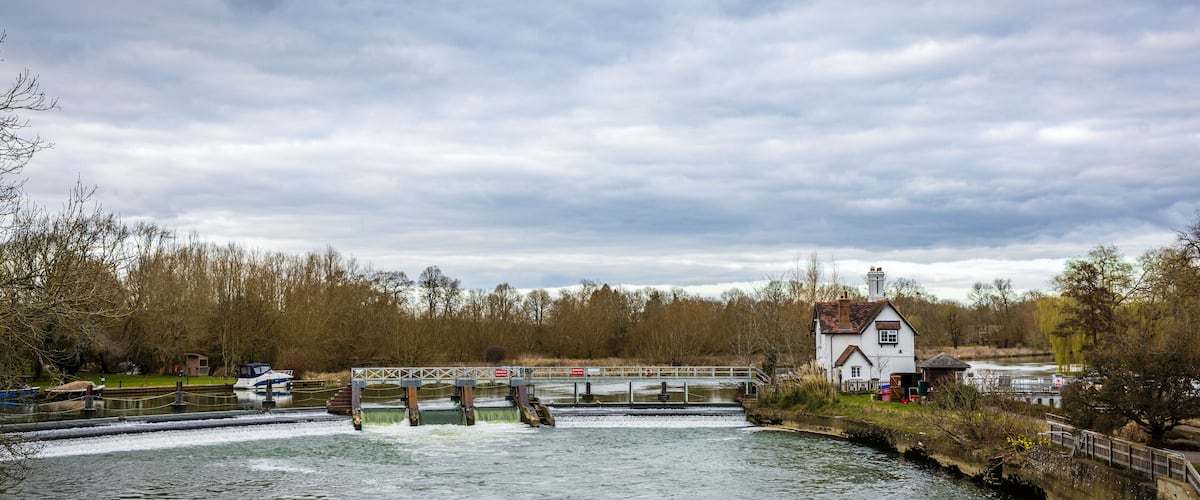 A river with a lock system, a white house, and trees in the background.