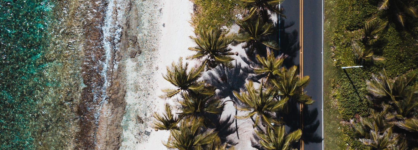 Vista aérea de una playa con agua turquesa cristalina, una orilla arenosa y palmeras bordeando la carretera.