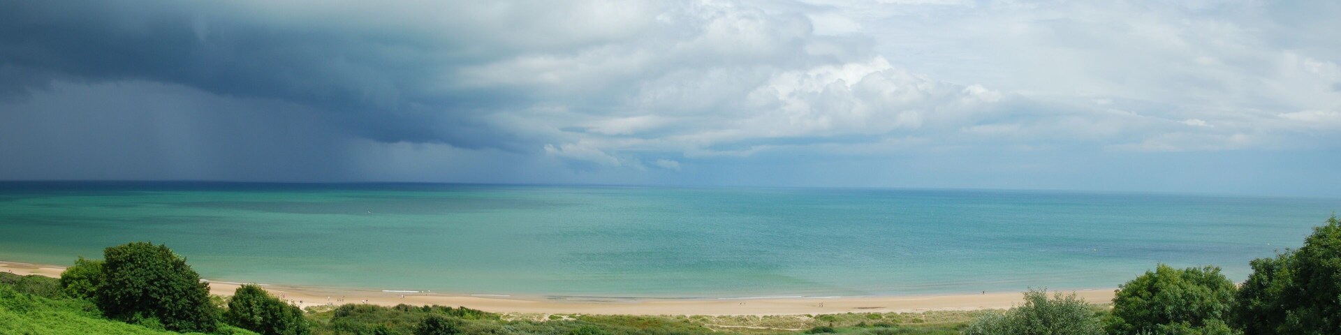 Uma praia com águas turquesa cristalinas, um areal e colinas verdejantes ao fundo.