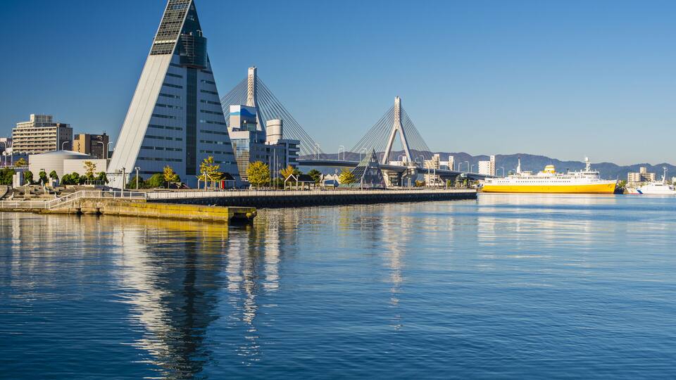 A modern cityscape with a distinctive triangular building and a cable-stayed bridge over a calm body of water.
