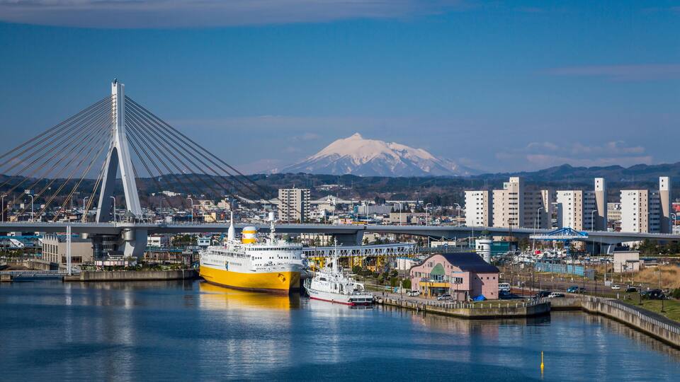 A yellow ferry docked at a port with a bridge and a snow-capped mountain in the background.