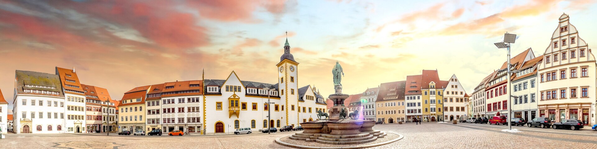 Una plaza adoquinada con edificios históricos, una destacada torre del reloj y una fuente con estatuas.