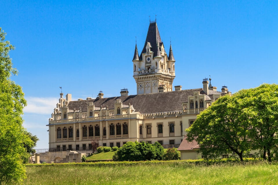 A large, ornate castle with a prominent clock tower and multiple spires, surrounded by lush greenery and a clear blue sky.