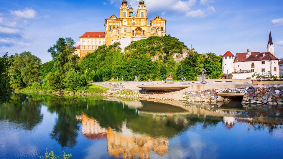 A castle on a hill with a reflection in the water, surrounded by greenery.