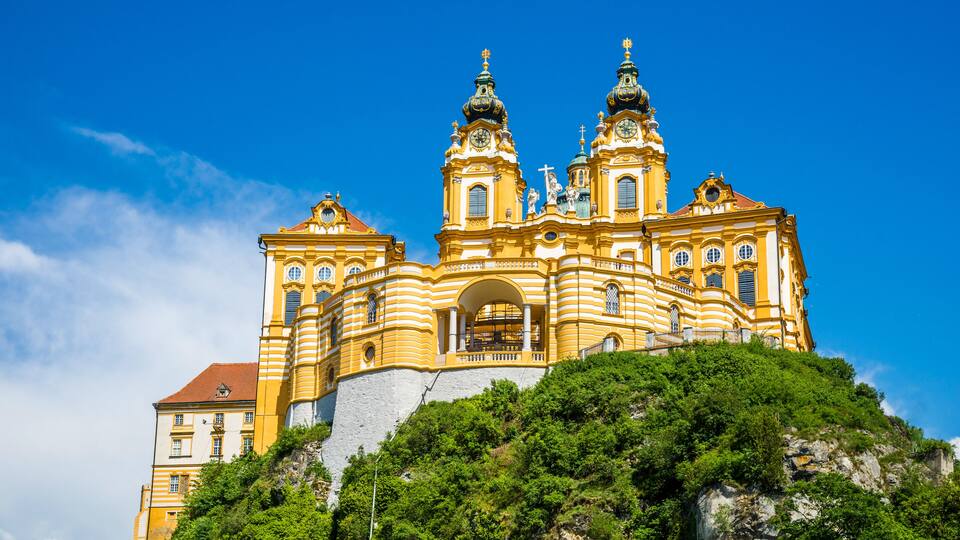 A large, ornate church with two towers and a bell tower, surrounded by lush greenery and perched on a hill.