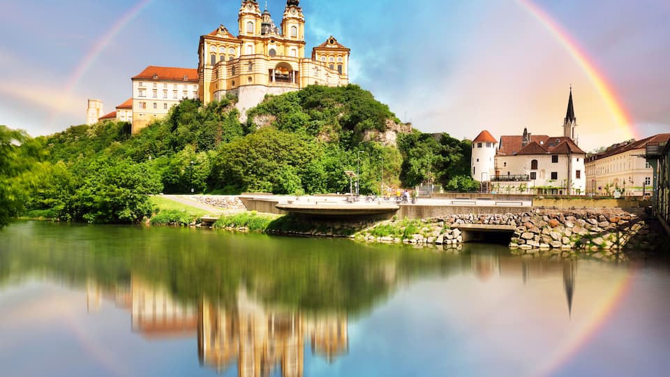 A cathedral with two towers, a rainbow, and a calm river reflecting the scene.