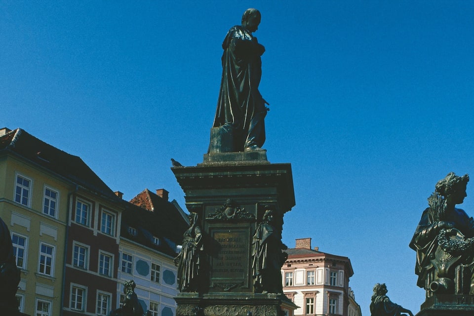 Estatua de una mujer con vestido sobre un pedestal con inscripciones, rodeada de edificios.