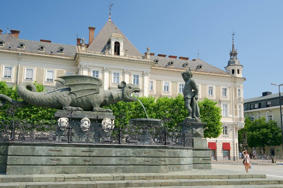 Una statua di un drago e di un uomo in una piazza con una fontana, di fronte a un edificio storico.