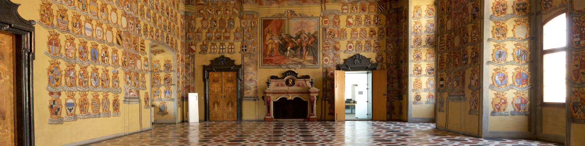 Un vaste hall doté de plafonds richement décorés, d’une grande fresque murale et de nombreuses armoiries sur les murs.
