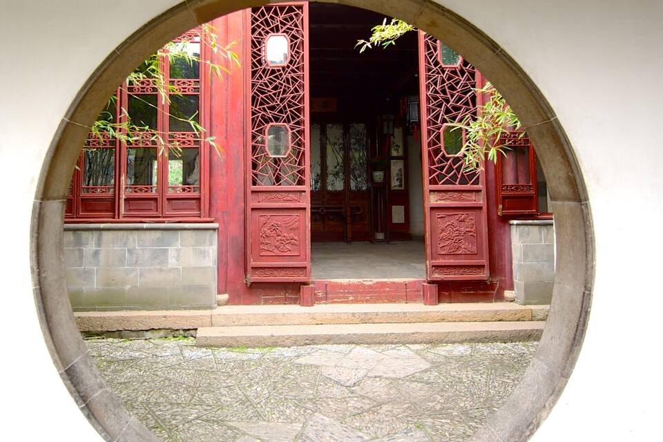 A traditional red gate with intricate designs, leading to a stone pathway and a building with wooden lattice windows.